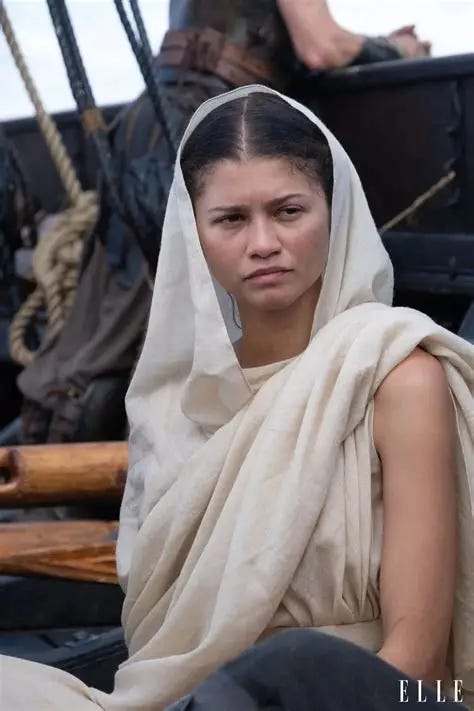 Zendaya: Retrato da atriz Zendaya caracterizada para uma produção cinematográfica, vestindo trajes beges com um manto sobre a cabeça em um cenário que remete a um barco antigo.