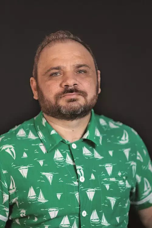 João Tordo: Retrato de um homem com barba vestindo uma camisa verde com estampa de barcos a vela sobre fundo escuro.