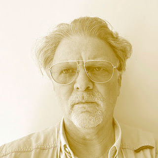 Eduardo Pitta: Retrato em tom sépia de um homem de meia-idade com óculos de aviador e barba curta, olhando para a câmera.