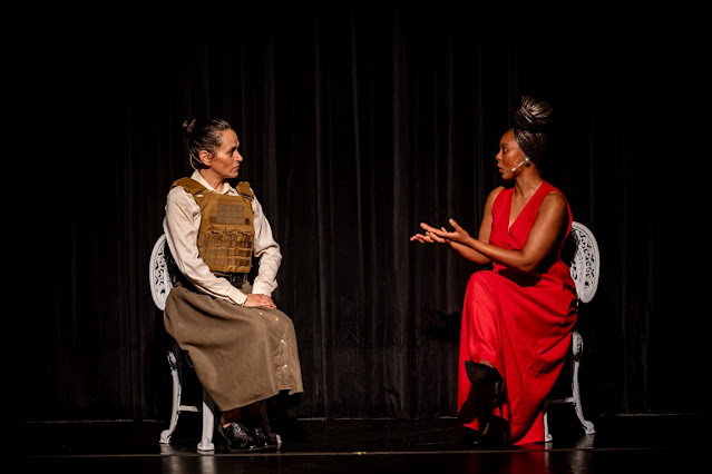 Duas atrizes em cena de teatro, uma vestindo um colete tático e outra um vestido longo vermelho, sentadas em cadeiras brancas contra um fundo preto.