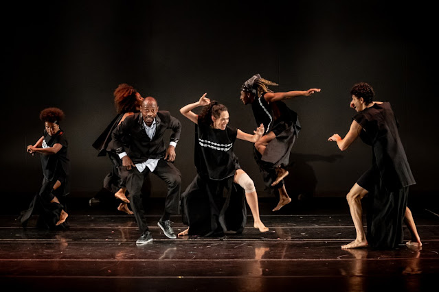Grupo de dançarinos negros realizando uma performance de dança contemporânea em um palco com fundo preto.
