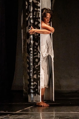 Uma mulher em cena teatral vestindo branco, posicionada entre elementos cenográficos verticais com padrões abstratos em preto e branco.