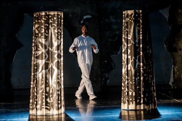 Um homem negro vestido de branco caminha em um palco escuro entre duas colunas translúcidas com padrões geométricos.