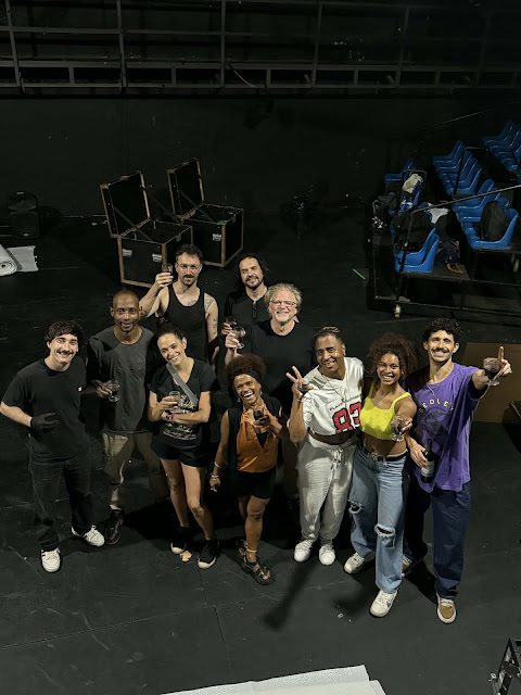 Um grupo de pessoas posa sorrindo em um palco de teatro ou estúdio, algumas segurando taças, possivelmente celebrando após uma produção.