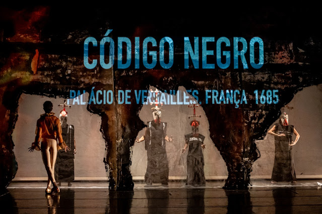 Uma cena teatral com atores mascarados e figurinos escuros sob o título 'Código Negro', mencionando o Palácio de Versalhes, França, 1685.