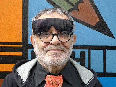 Fernando Relvas: Retrato frontal de um homem idoso com barba grisalha, usando óculos de grau redondos e um protetor solar/viseira escura sobre a testa, posando em frente a um mural colorido.