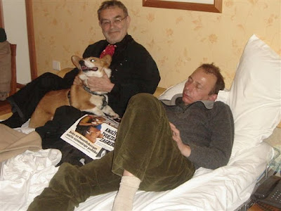 Houellebecq: Dois homens em uma cama, um sentado segurando um cachorro Corgi e outro deitado ao lado, com um jornal sobre o colo.