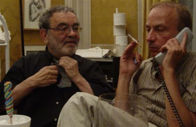 Fernando Gomes e José Eduardo Agualusa: Dois homens de meia-idade em uma conversa informal; um ajusta a gravata e o outro segura um cigarro enquanto fala ao telefone.