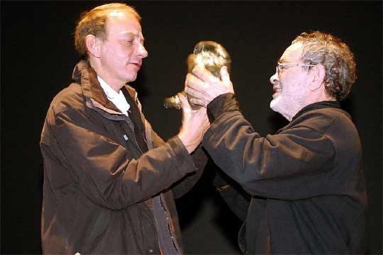 Eduardo Prado Coelho: Dois homens de perfil segurando conjuntamente uma escultura dourada em formato de punho ou pé sobre um fundo preto.