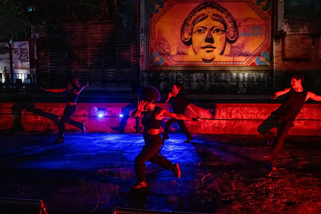 Pessoas realizando uma performance de dança contemporânea em um ambiente externo noturno com iluminação dramática em tons de azul e vermelho, sob um painel artístico.