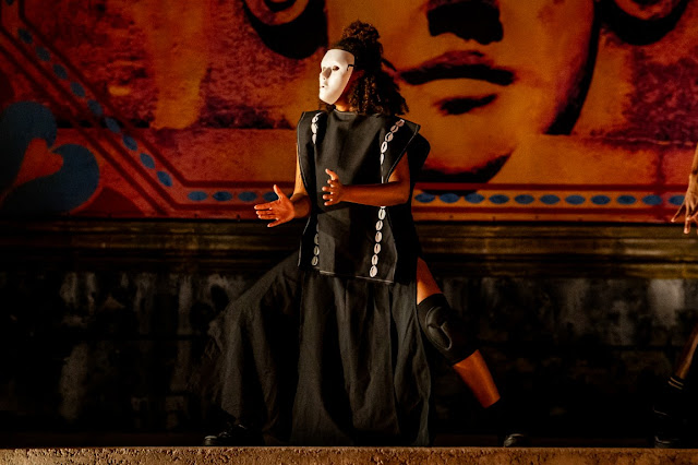 Uma pessoa performando com uma máscara branca e vestimentas pretas em frente a um painel artístico de tons quentes.