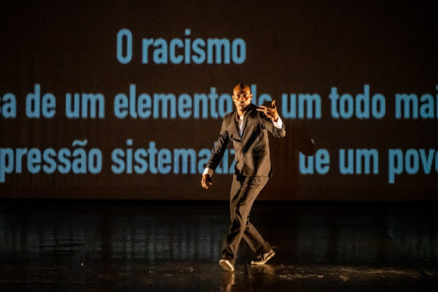 Mário Coelho: Homem negro em traje formal realizando uma performance artística ou dança em um palco, com projeção de texto sobre racismo ao fundo.