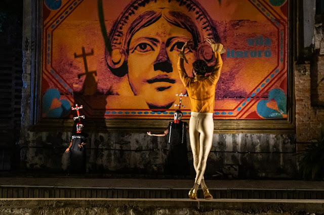 Performance artística ou teatral em frente a um grande mural com o rosto de uma mulher e o texto 'Vila Itororó'.