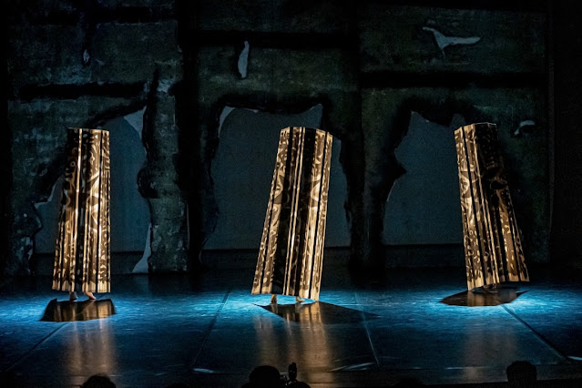 Fotografia de uma performance teatral ou de dança contemporânea, apresentando três figuras cobertas por estruturas cilíndricas padronizadas sob iluminação de palco.