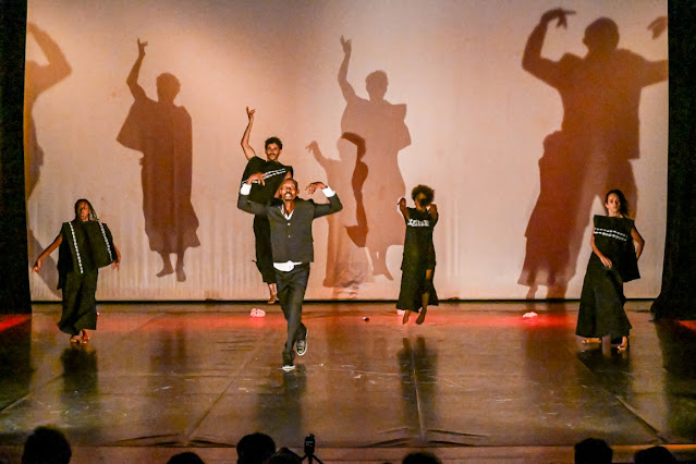 Uma apresentação artística de dança contemporânea em um palco, com bailarinos negros performando em frente a um telão que projeta suas sombras ampliadas.