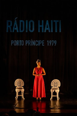 Uma mulher em um vestido vermelho em pé no palco entre duas cadeiras brancas ornamentadas, com o texto 'RÁDIO HAITI PORTO PRÍNCIPE 1979' projetado ao fundo.