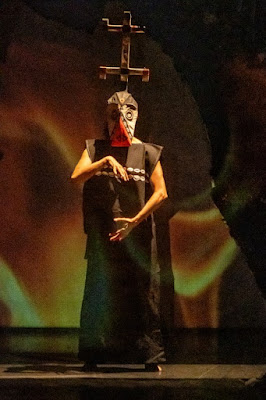 Uma pessoa vestida com trajes e máscara performática em um palco, possivelmente uma cena de teatro ou dança com influências culturais africanas.