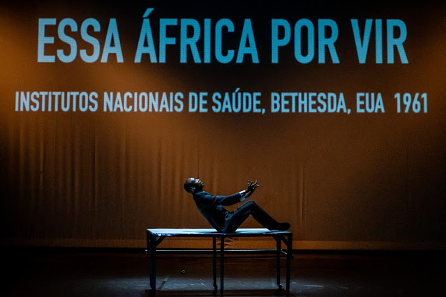 Um ator em performance teatral sentado sobre uma mesa, com projeção de texto ao fundo mencionando 'ESSA ÁFRICA POR VIR' e 'INSTITUTOS NACIONAIS DE SAÚDE, BETHESDA, EUA 1961'.