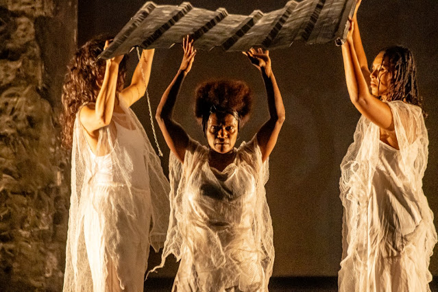 Três mulheres negras em cena de teatro, vestindo roupas brancas diáfanas e segurando uma estrutura de madeira sobre as cabeças.