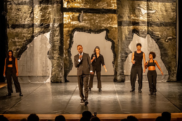 Cinco artistas em um palco de teatro com um cenário abstrato ao fundo. O homem ao centro veste um terno e segura um microfone, enquanto os outros quatro estão em trajes pretos casuais ou esportivos.