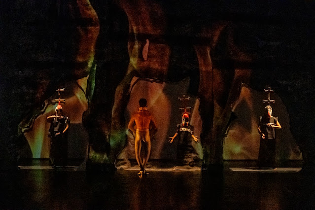 Performers on a theatrical stage with elaborate costumes and a backdrop featuring abstract organic shapes and projections.