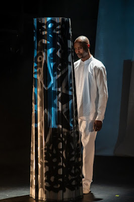 Um homem negro vestido de branco em um palco, posicionado atrás de uma coluna decorativa com padrões vazados.