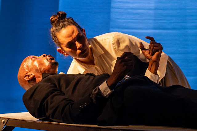 Uma cena de performance teatral onde um homem negro em um terno escuro está deitado em uma plataforma enquanto uma mulher com cabelo preso em um coque e camisa branca o observa intensamente.