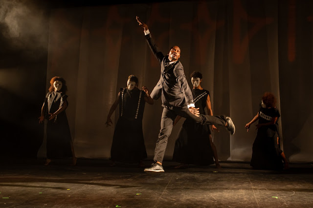 Performers em um palco de teatro, com um homem em destaque saltando e outros quatro ao fundo usando máscaras brancas e trajes escuros.