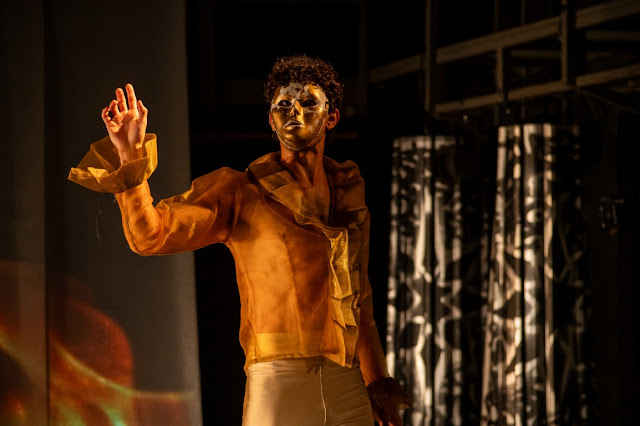 Um ator no palco usando uma máscara dourada, camisa amarela translúcida com babados e calças brancas, com a mão levantada em um gesto dramático.