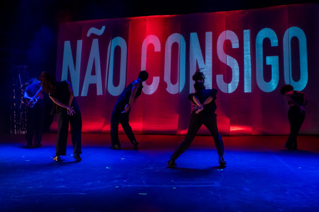 Performers on a stage with blue lighting, in front of a backdrop with the text 'NÃO CONSIGO' illuminated in red and white.