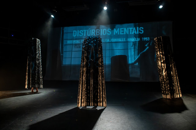 Uma cena de palco teatral com três estruturas altas e vazadas sob iluminação dramática, com uma projeção ao fundo contendo o texto 'DISTÚRBIOS MENTAIS' e referências a um hospital em 1953.