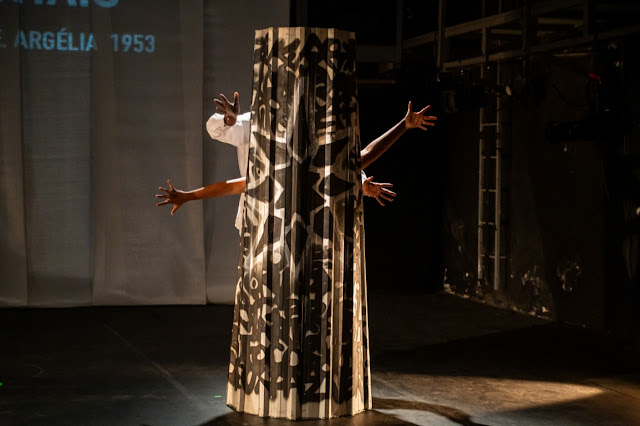 Uma performance artística ou peça teatral no palco, onde braços humanos emergem de uma estrutura cilíndrica decorada com padrões pretos.