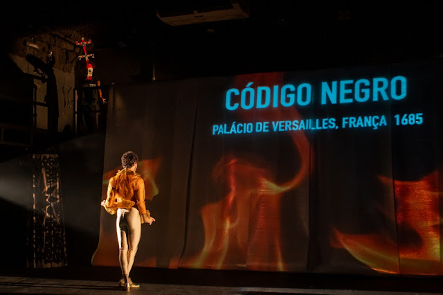Uma fotografia de uma performance teatral onde um ator está de costas para a câmera, observando uma projeção que diz 'CÓDIGO NEGRO - PALÁCIO DE VERSAILLES, FRANÇA 1685' sobre um fundo de chamas.