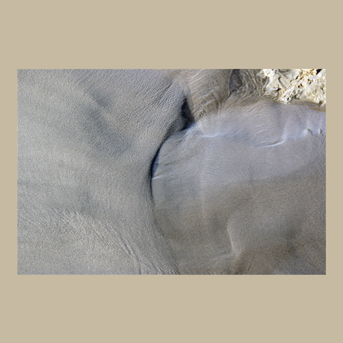 Uma fotografia abstrata de areia molhada com padrões formados pela água e uma rocha no canto superior.