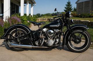 Uma motocicleta Harley-Davidson clássica de cor preta estacionada em uma calçada em frente a uma casa.