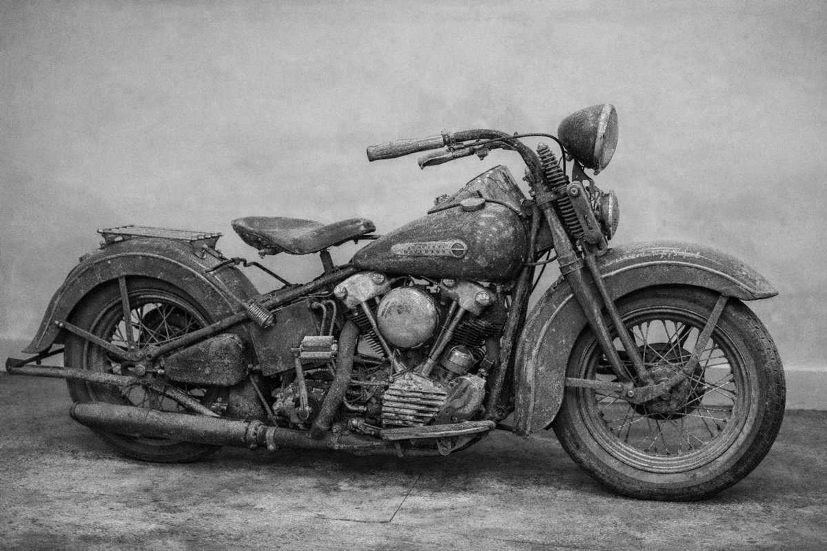 Uma fotografia em preto e branco de uma motocicleta antiga e empoeirada, possivelmente uma Harley-Davidson clássica, vista de perfil.