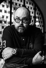Carlos Ruiz Zafón: Retrato em preto e branco de um homem com óculos redondos, barba e braços cruzados.