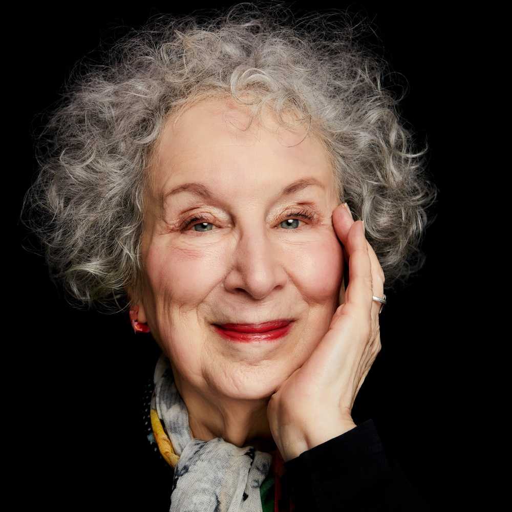 Margaret Atwood: Retrato de close-up de uma mulher idosa com cabelos grisalhos cacheados, sorrindo com a mão no rosto sobre um fundo preto.