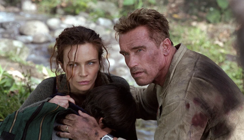 Arnold Schwarzenegger, Francesca Neri: Uma cena do filme 'Collateral Damage' (Efeito Colateral) mostrando um homem e uma mulher agachados em uma área externa, protegendo uma criança.