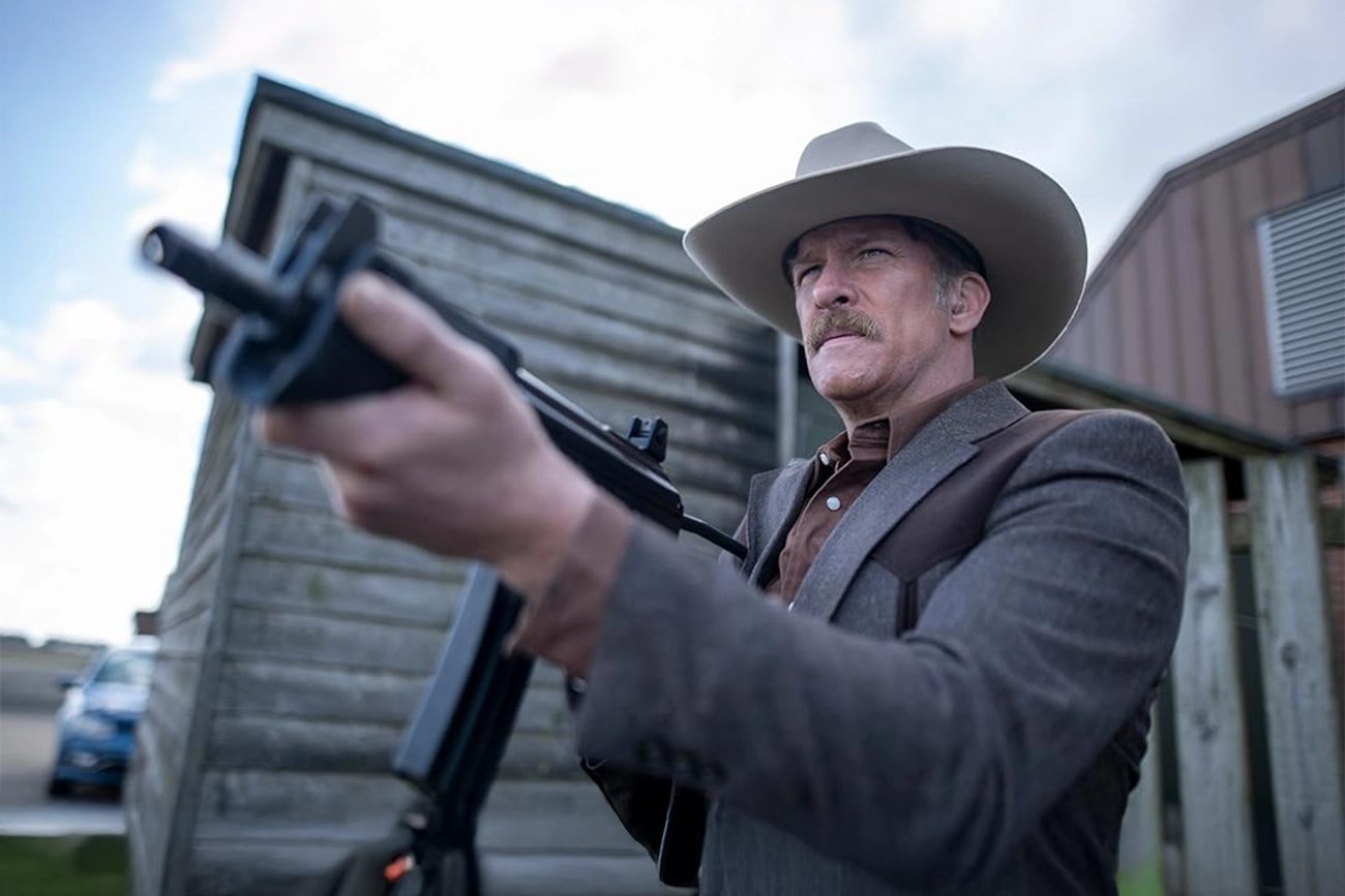 Barry Pepper: Homem de chapéu de cowboy segurando uma arma automática em frente a um galpão de madeira.