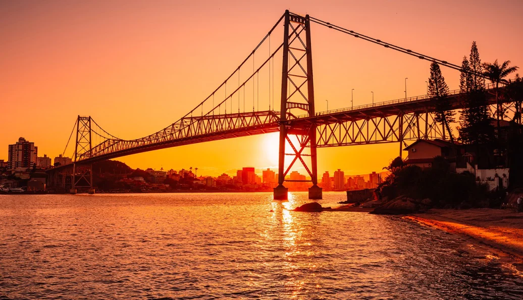 Vista da Ponte Hercílio Luz em Florianópolis durante o pôr do sol, com a silhueta da ponte e da cidade ao fundo.