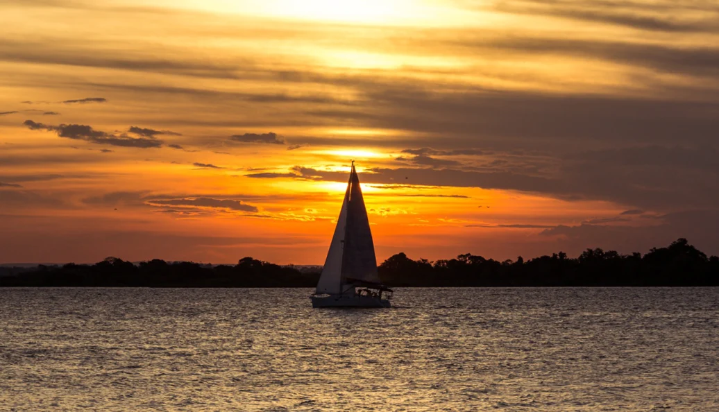 Um veleiro navegando em águas calmas durante um pôr do sol dourado, com silhuetas de árvores ao fundo.
