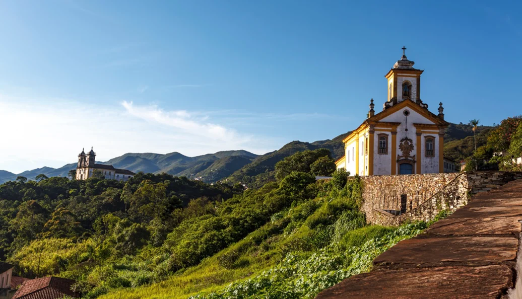 unknown: Uma fotografia panorâmica mostrando igrejas históricas de estilo colonial em Ouro Preto, Minas Gerais, situadas em colinas verdes sob um céu azul claro.
