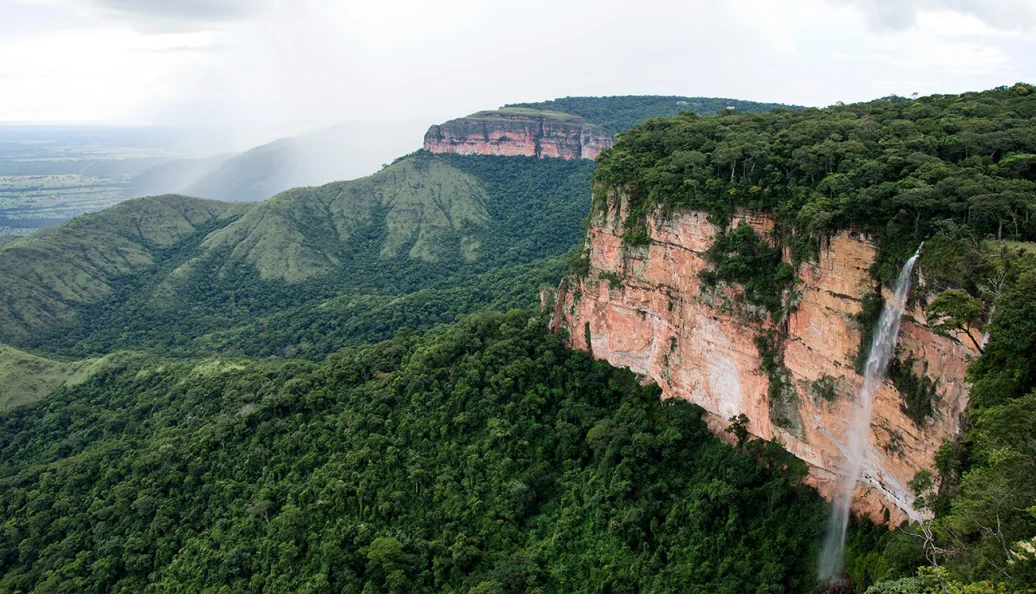 Uma paisagem panorâmica da Chapada dos Guimarães, mostrando paredões de arenito, floresta densa e a cachoeira Véu de Noiva.