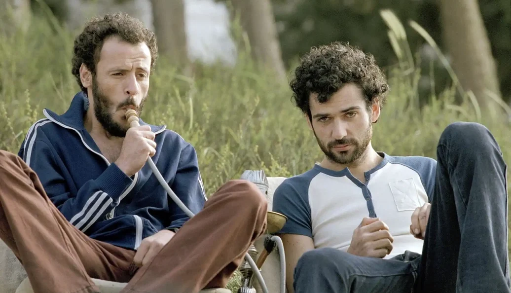Alboum e Mounir Margoum: Dois homens sentados ao ar livre, um deles fumando um narguilé.