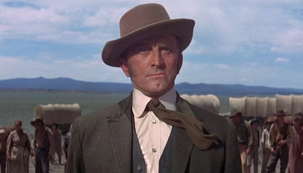 Kirk Douglas: Retrato de Kirk Douglas em um cenário de filme de faroeste, caracterizado com chapéu e roupas de época à frente de uma caravana.