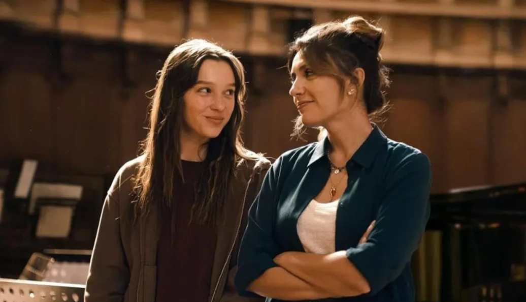 unknown: Duas mulheres em um ambiente interno, possivelmente um estúdio ou teatro, olhando uma para a outra e sorrindo.