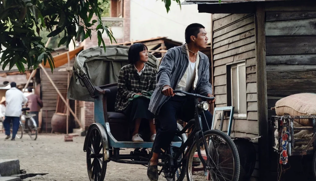 Tony Yang, Zhou Dongyu: Uma cena de filme mostrando um homem puxando uma mulher em um riquixá de bicicleta em uma vila rural.