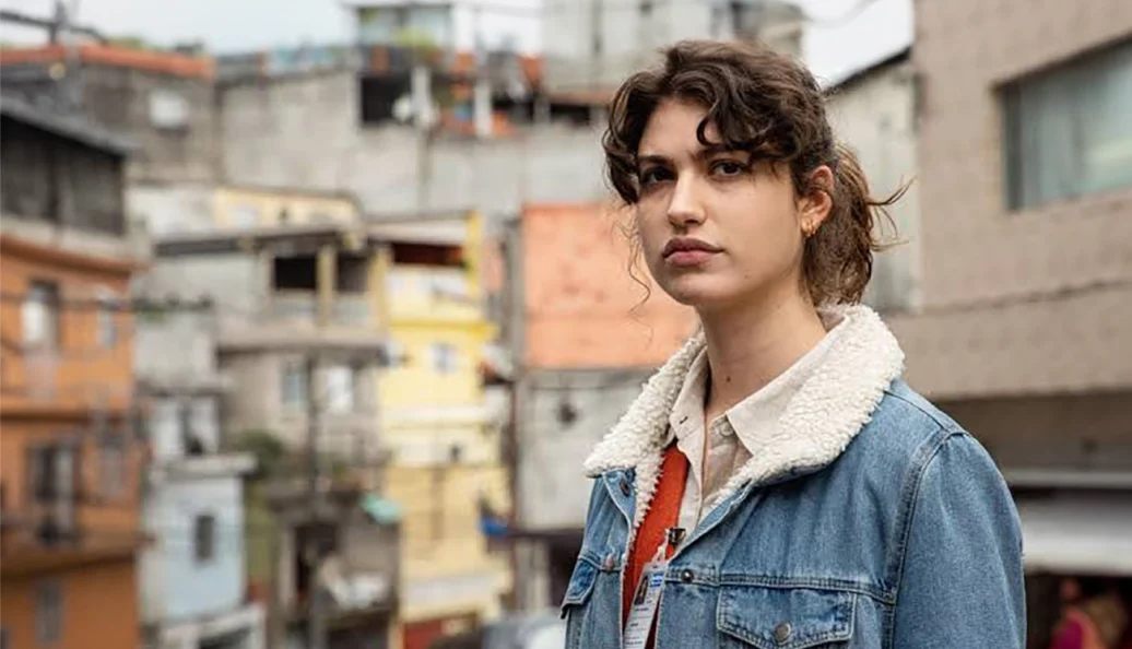 Giovana Grigio: Uma mulher jovem com cabelos cacheados e jaqueta jeans em frente a uma área residencial urbana densa, possivelmente uma favela.