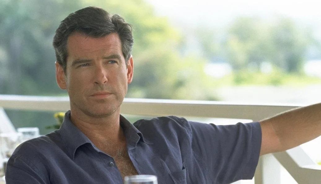 Pierce Brosnan: Retrato de um homem de meia-idade vestindo uma camisa social azul, sentado em um ambiente externo ensolarado.